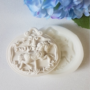 Puede incluir: Un molde de silicona blanco para hacer un adorno de caballo de carrusel. El molde está en dos piezas, con el caballo y su marco decorativo en una pieza y la base en la otra. El molde se muestra sobre un fondo blanco.