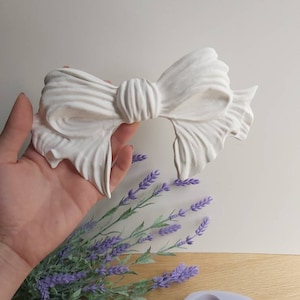 Pode incluir: Forma de silicone branca para fazer um grande laço decorativo. A forma é mostrada com um laço acabado feito de resina branca.