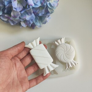 Puede incluir: Un molde de silicona blanco para hacer caramelos. El molde tiene dos cavidades, una con forma de bastón de caramelo y la otra con forma de caramelo redondo con un diseño de espiral.