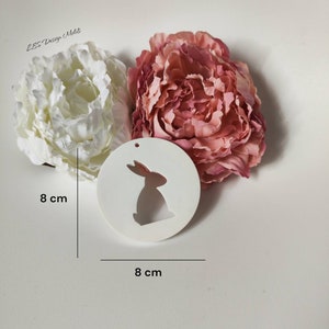 May include: A white, circular tag with a cut-out bunny design, measuring 8 cm in diameter, is displayed with a white and a pink peony. The tag has a small hole at the top. The text "LBS Design Molit" is in the upper left corner.