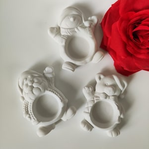 Pode incluir: Três argolas de guardanapo de Natal em cerâmica branca. Uma argolla apresenta um boneco de neve, outra o Papai Noel e a terceira um urso com uma echarpe e um laço de gravata.