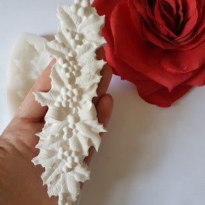 Könnte beinhalten: Weiße Silikonform einer Stechpalmen-Girlande mit Beeren. Die Form wird in einer Hand vor einem weißen Hintergrund gehalten.