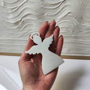 Può includere: Ciondolo angelo in legno bianco con aureola. L'angelo tiene le mani giunte davanti al petto.