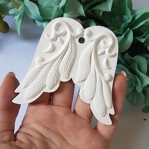 May include: White ceramic angel wings with a detailed feather design. The wings are held in a hand.
