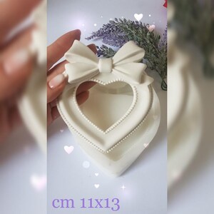 May include: White ceramic heart-shaped picture frame with a bow on top. The frame measures 11x13 cm.