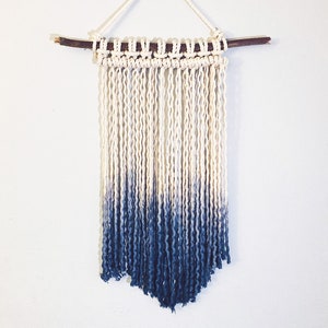 Puede incluir: Un tapiz de macramé de color blanco y azul degradado con una barra de madera en la parte superior.