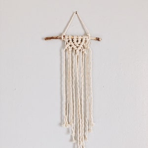 May include: A white macrame wall hanging with a natural wood branch at the top. The macrame is made with a simple knot pattern and has a fringe at the bottom.