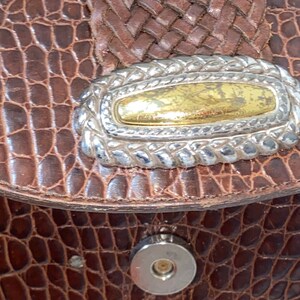 May include: Close-up of a brown leather purse with a silver and gold decorative accent. The purse has a crocodile-embossed texture.