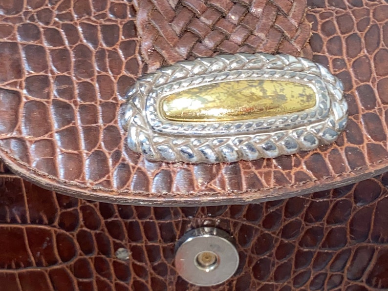 May include: Close-up of a brown leather purse with a silver and gold decorative accent. The purse has a crocodile-embossed texture.
