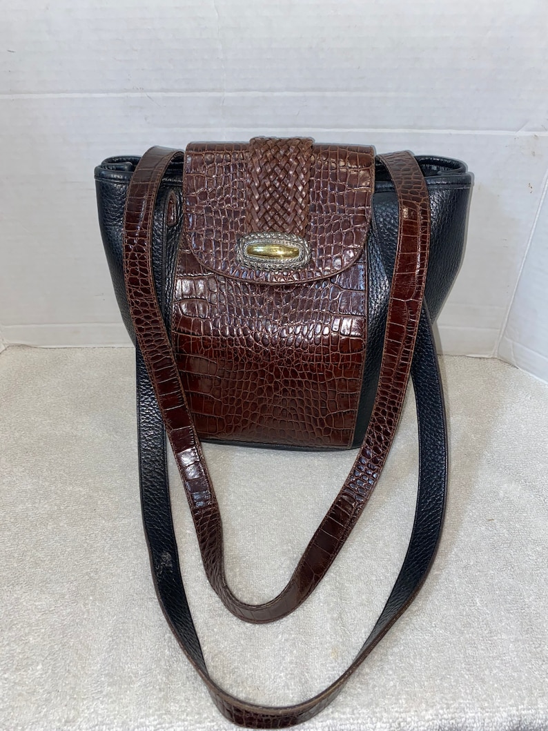 May include: A brown and black leather handbag with a crocodile embossed pattern. The bag has a flap closure with a decorative metal clasp. The bag has a long adjustable strap.