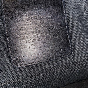 May include: Close-up of a black leather tag with white text that reads "The bags are individually crafted from the finest. Any variations in surface are part of the natural beauty and character of the leather with time. No. 84102"