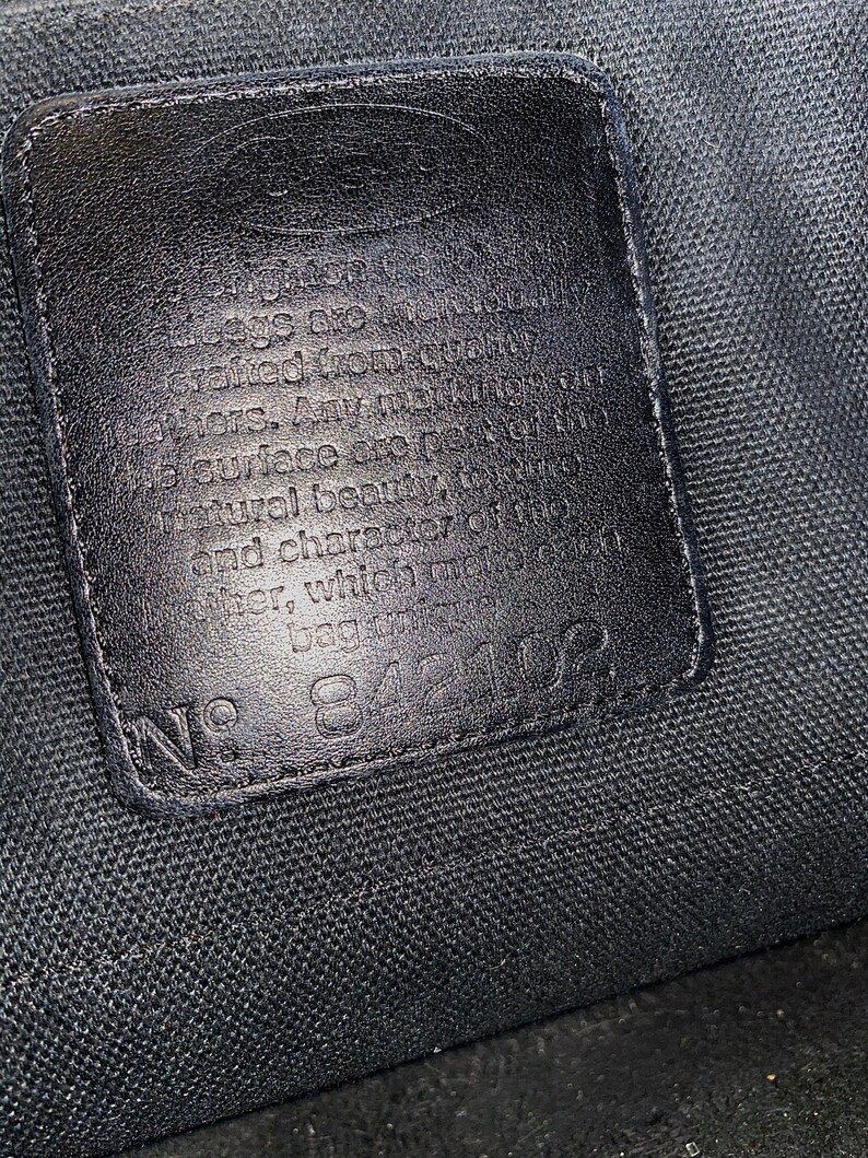 May include: Close-up of a black leather tag with white text that reads "The bags are individually crafted from the finest. Any variations in surface are part of the natural beauty and character of the leather with time. No. 84102"