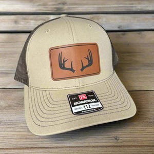 Pode incluir: Um boné de caminhoneiro bege e marrom com um remendo de couro marrom que apresenta uma silhueta de uma galhada de veado. O boné tem uma malha preta nas costas e o texto "EST. 1970 RICHARDSON. ORIGINAL 112 TRUCKER ADJUSTABLE" nas costas.