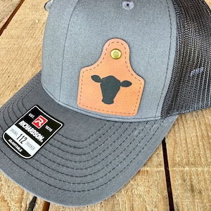 May include: A gray and black trucker hat with a brown leather patch featuring a black silhouette of a cow's head. The hat has an adjustable strap and the text "Richardson" and "Original 112 Trucker" is visible on the strap.