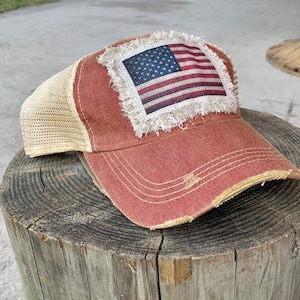 May include: A red and beige trucker hat with a distressed look. The hat features a frayed patch with an American flag design.