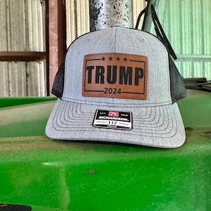 May include: A grey and black trucker hat with a brown leather patch that says "TRUMP - 2024". The hat is sitting on a green surface.