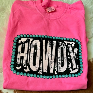 May include: Pink t-shirt with a turquoise and white border around the word "HOWDY" in black and white with a distressed look.
