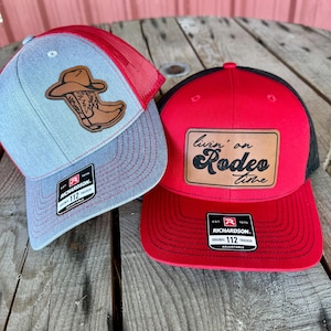 Puede incluir: Dos gorras de camionero con temática de vaquero. Una es azul claro con un parche de cuero marrón con una bota y un sombrero de vaquero. La otra es roja con una parte trasera de malla negra y un parche de cuero que dice "Livin' on Rodeo Time".