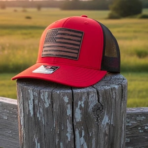 May include: A red baseball cap with a black mesh back and a brown leather patch featuring a distressed American flag. The cap is sitting on a wooden post in an outdoor setting.