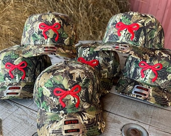May include: A collection of camouflage baseball caps with a red bow embroidered on the front. The caps are in various positions, showcasing the design and the "OTTO" brand label. The hats are on a wooden surface.