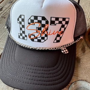 May include: A black and white checkered trucker hat with the number 137 and the word "Shine" in orange. The hat has a black mesh back and a silver chain with a checkered heart charm.