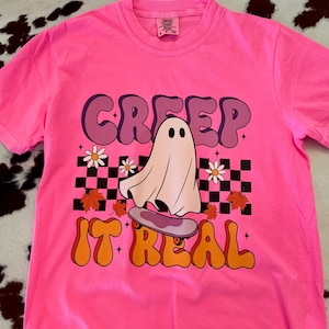 May include: A bright pink short-sleeve t-shirt featuring a graphic of a white ghost on a skateboard. The design includes a black and white checkered pattern, orange autumn leaves, white daisies, and the text CREEP IT REAL in orange.