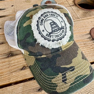 Op de afbeelding: Een camouflage truckercap met een wit mesh achterpaneel. De pet heeft een distressed, witte patch met een zwart-witte afbeelding van een slang met de tekst "Come and Take It Don't Tread On Me".