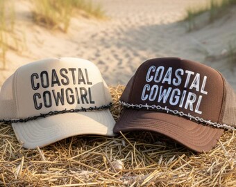 Könnte beinhalten: Zwei Trucker-Hüte mit dem Text "COASTAL COWGIRL". Einer ist beige mit einem schwarzen Stacheldraht-Design, der andere ist braun mit einem weißen Stacheldraht-Design. Die Hüte liegen auf Heu mit Strand und Meer im Hintergrund.
