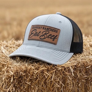 May include: A gray and black trucker hat with the text "SUPPORT RANCHERS Eat Beef" on a brown leather patch. The hat is sitting on a bale of hay.