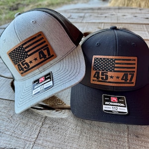 Könnte beinhalten: Zwei Trucker-Caps mit schwarzem Netzrücken. Die Caps sind grau und schwarz. Beide Caps haben ein braunes Leder-Patch mit einer amerikanischen Flagge und dem Text "45**47". Die Caps haben auch ein kleines Etikett mit dem Text "RICHARDSON. ORIGINAL 112 TRUCKER YOUTH".