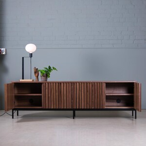 May include: A dark brown wooden cabinet with vertical grooves and black metal legs. The cabinet has two doors that are open, revealing shelves inside. A small plant and a decorative hand are on top of the cabinet.