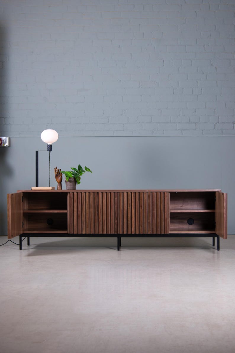 May include: A dark brown wooden cabinet with vertical grooves and black metal legs. The cabinet has two doors that are open, revealing shelves inside. A small plant and a decorative hand are on top of the cabinet.