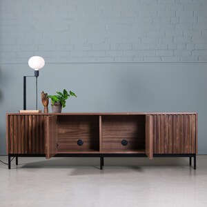May include: A dark wood media console with three doors, two of which are open, revealing empty shelves. The console has black metal legs and is sitting on a light gray floor.