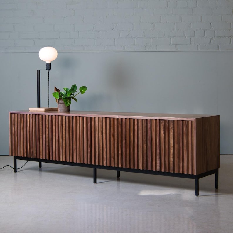 May include: A long, dark brown wooden media console with vertical grooves and black metal legs. The console has four doors and is sitting on a light gray floor.