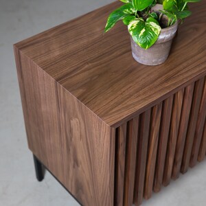 May include: A wooden cabinet with a slatted front and black legs. A potted plant with green leaves sits on top of the cabinet.