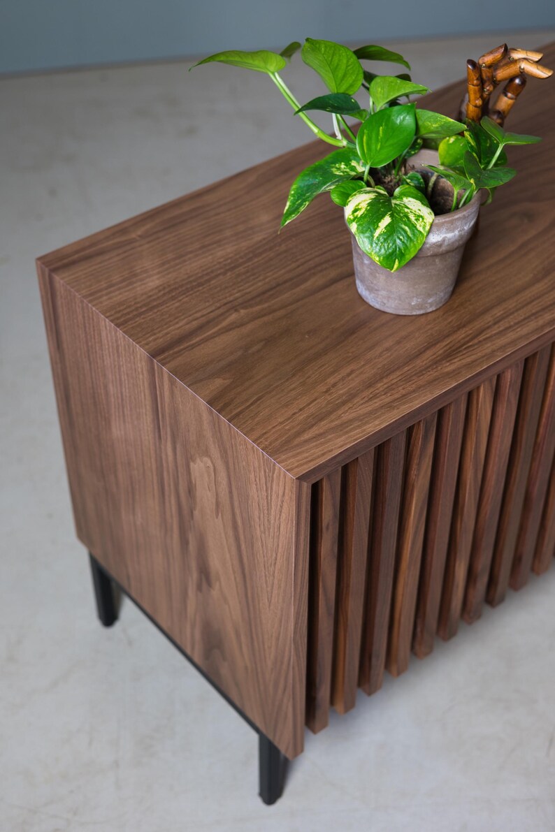 May include: A wooden cabinet with a slatted front and black legs. A potted plant with green leaves sits on top of the cabinet.