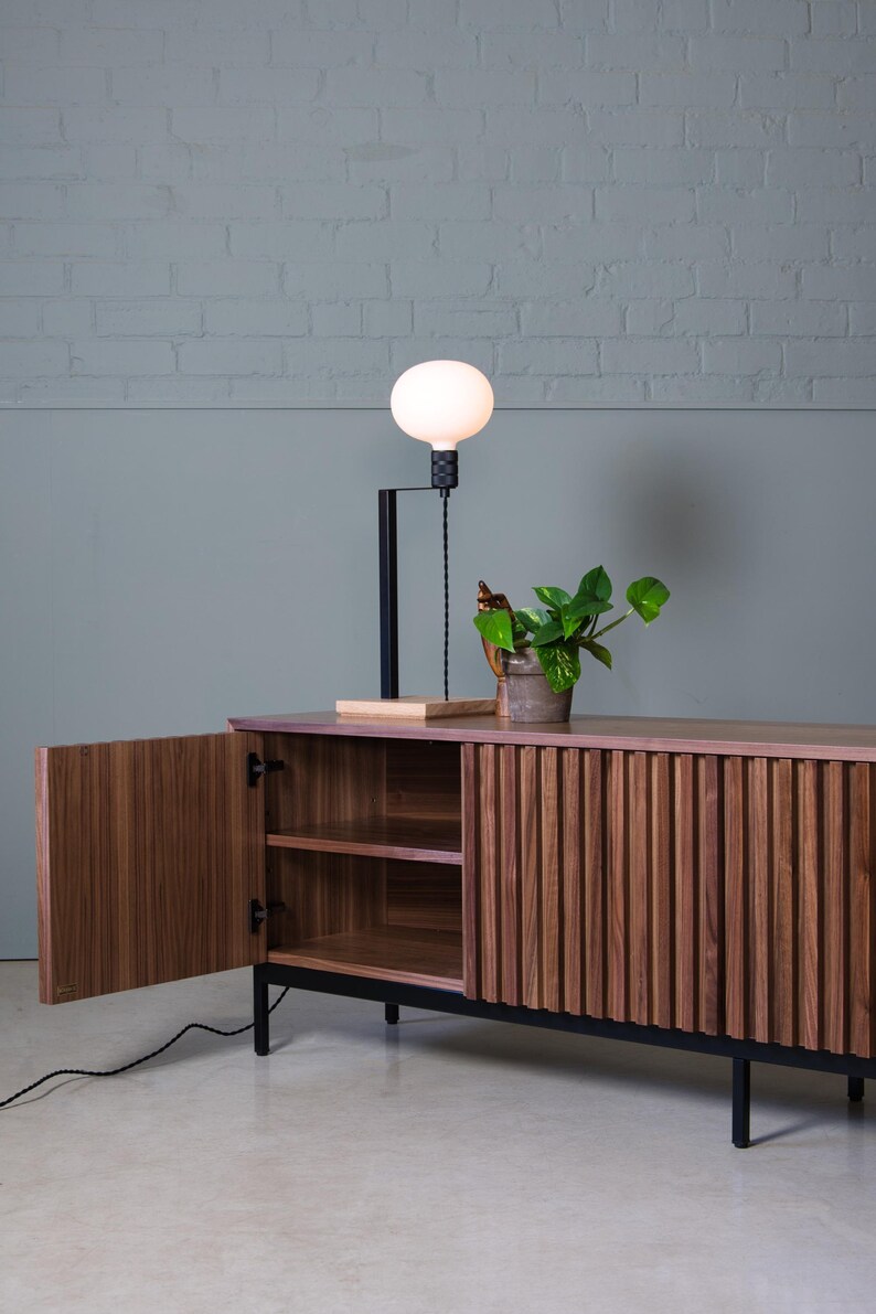 May include: A dark wood cabinet with two doors and a shelf inside. The cabinet has a black metal frame and legs. A small plant is sitting on top of the cabinet. A white globe lamp is on the wall behind the cabinet.