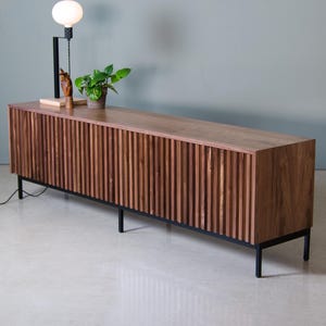 May include: A long, dark wood media console with a slatted front and black metal legs. The console is sitting on a white tiled floor.
