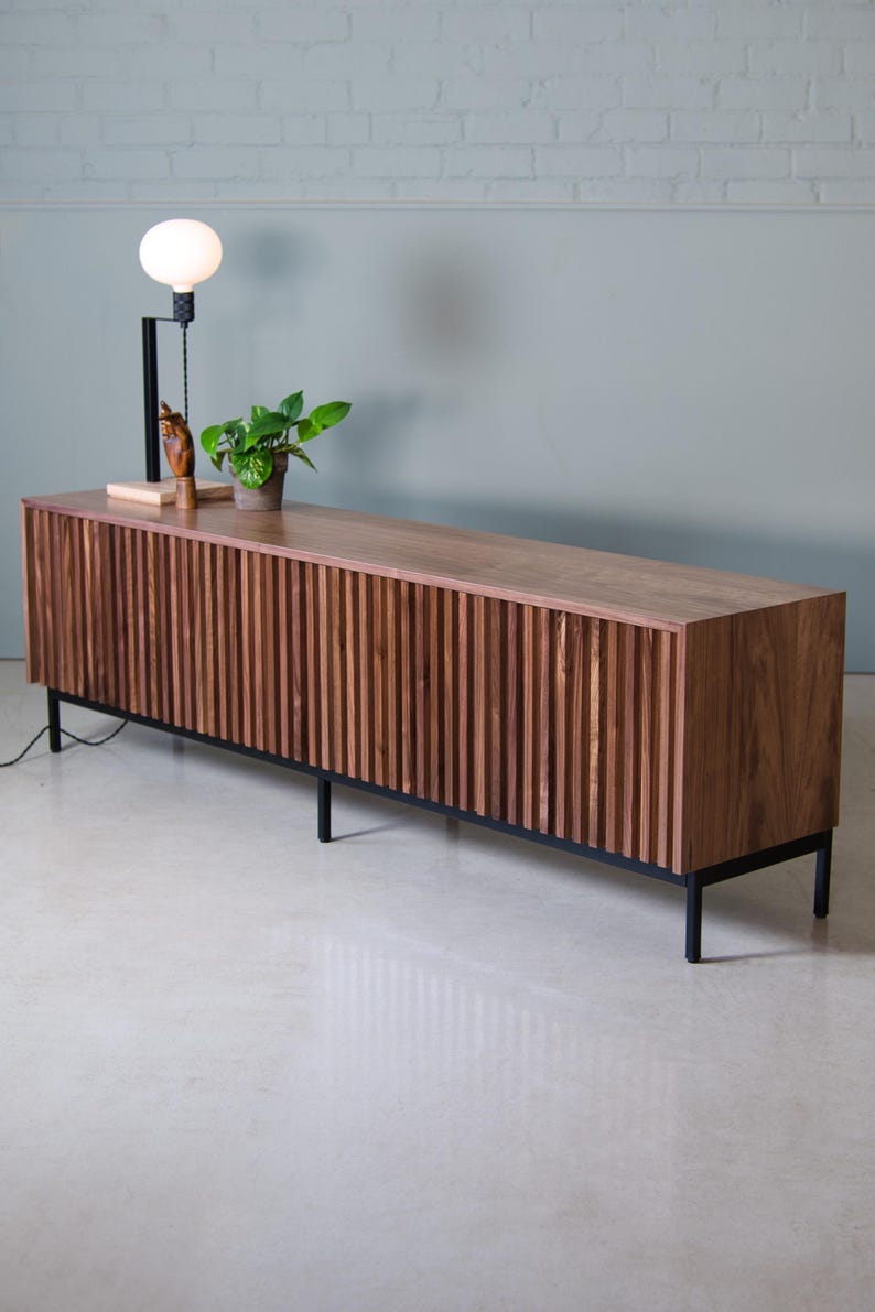 May include: A long, dark wood media console with a slatted front and black metal legs. The console is sitting on a white tiled floor.