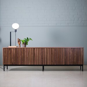 May include: A long, dark brown wooden cabinet with vertical slats and black metal legs. The cabinet is sitting on a light gray concrete floor in front of a white brick wall.