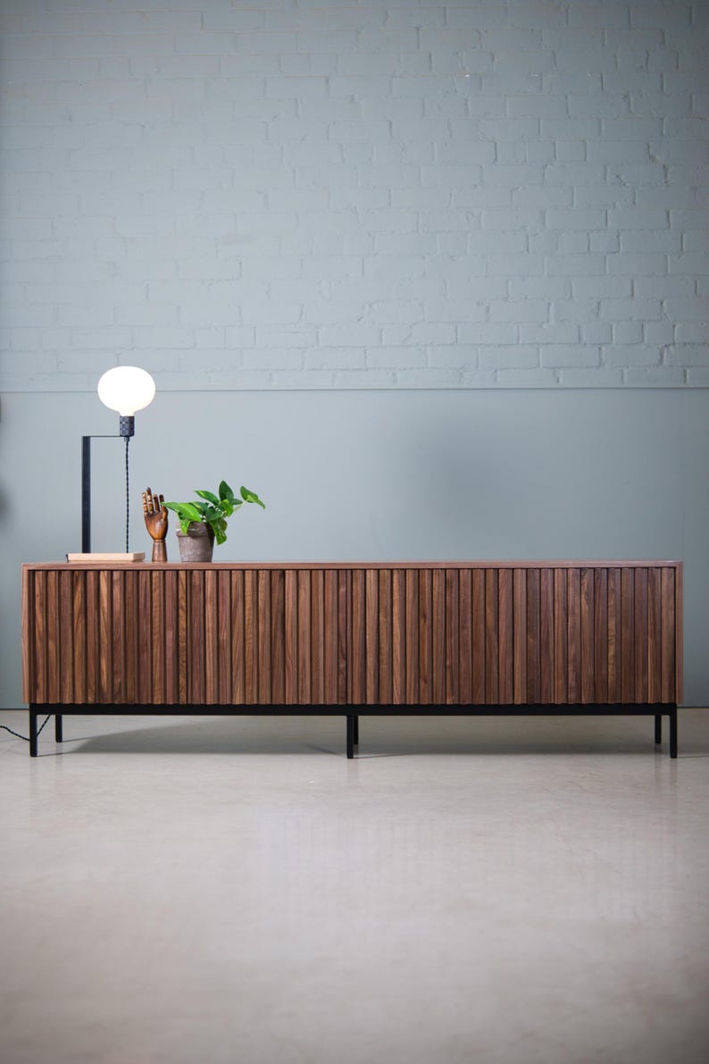 May include: A long, dark brown wooden cabinet with vertical slats and black metal legs. The cabinet is sitting on a light gray concrete floor in front of a white brick wall.