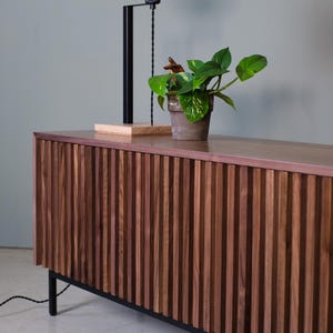 May include: A dark wood cabinet with vertical grooves and black metal legs. A plant in a terracotta pot sits on top of the cabinet, and a black lamp with a white globe is behind the plant.