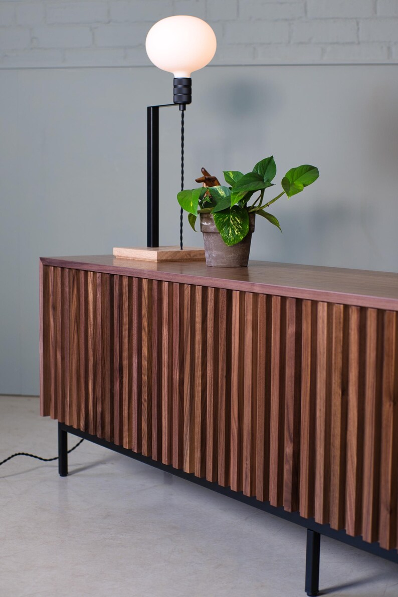 May include: A dark wood cabinet with vertical grooves and black metal legs. A plant in a terracotta pot sits on top of the cabinet, and a black lamp with a white globe is behind the plant.
