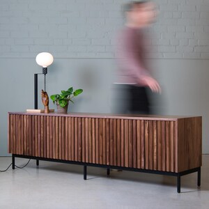 May include: A dark wood media console with vertical grooves and black metal legs. The console has two doors and is sitting on a light gray floor.