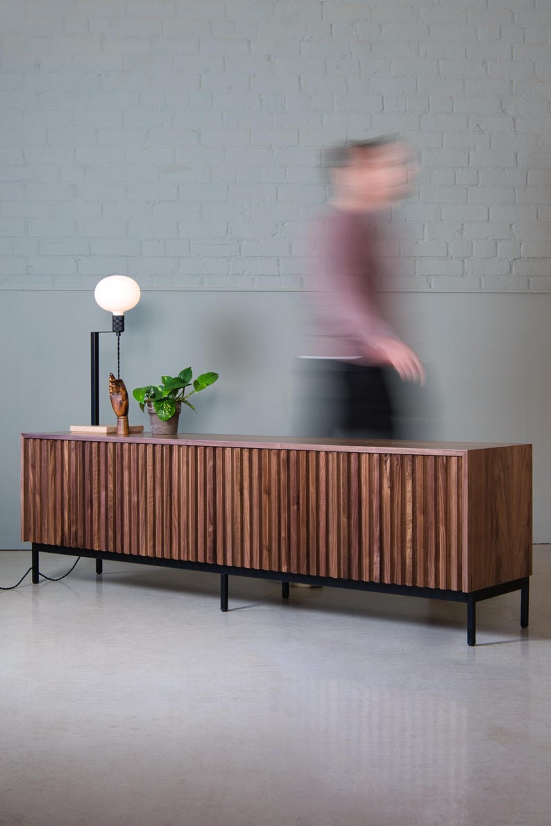 May include: A dark wood media console with vertical grooves and black metal legs. The console has two doors and is sitting on a light gray floor.