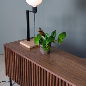May include: A wooden cabinet with vertical slats, a plant in a pot, and a black lamp with a white globe on top.