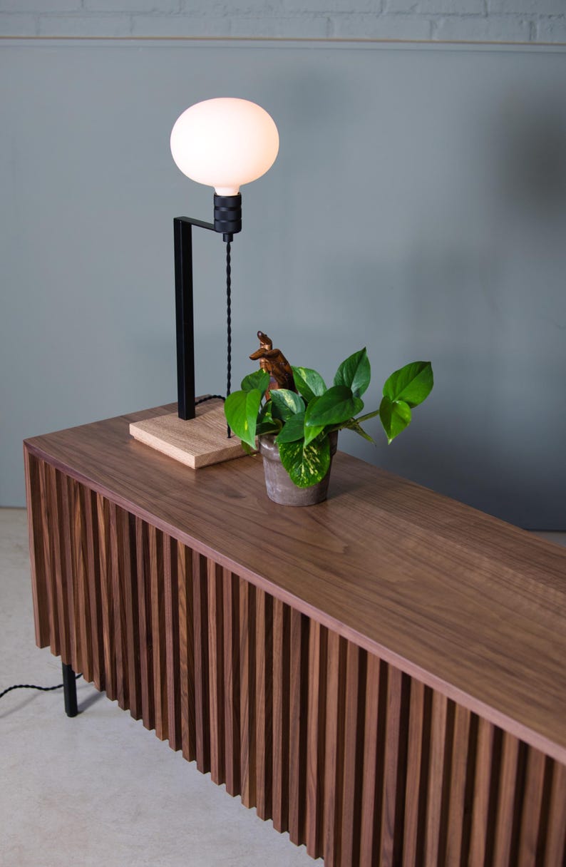 May include: A wooden cabinet with vertical slats, a plant in a pot, and a black lamp with a white globe on top.