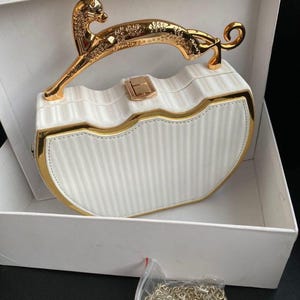 Op de afbeelding: Witte mini-tas met gouden details, waaronder een decoratieve handgreep met een dierenmotief. De tas wordt gepresenteerd in een witte doos, met een gouden ketting in een plastic zak. De tekst "Jm Mini bag White" is zichtbaar.
