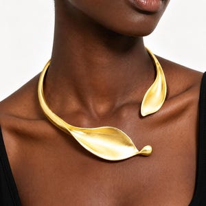 May include: A gold-toned metal choker necklace featuring an open-front design. The piece is shaped like two smooth, elongated, curved leaves that taper to rounded points, resting against the collarbone with a matte, reflective finish.
