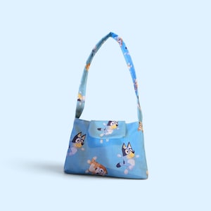 May include: A light blue shoulder bag featuring a repeating pattern of cartoon blue and orange dogs. The bag has a single shoulder strap and a rectangular body with a flap closure, showcasing multiple illustrations of the characters in various poses.